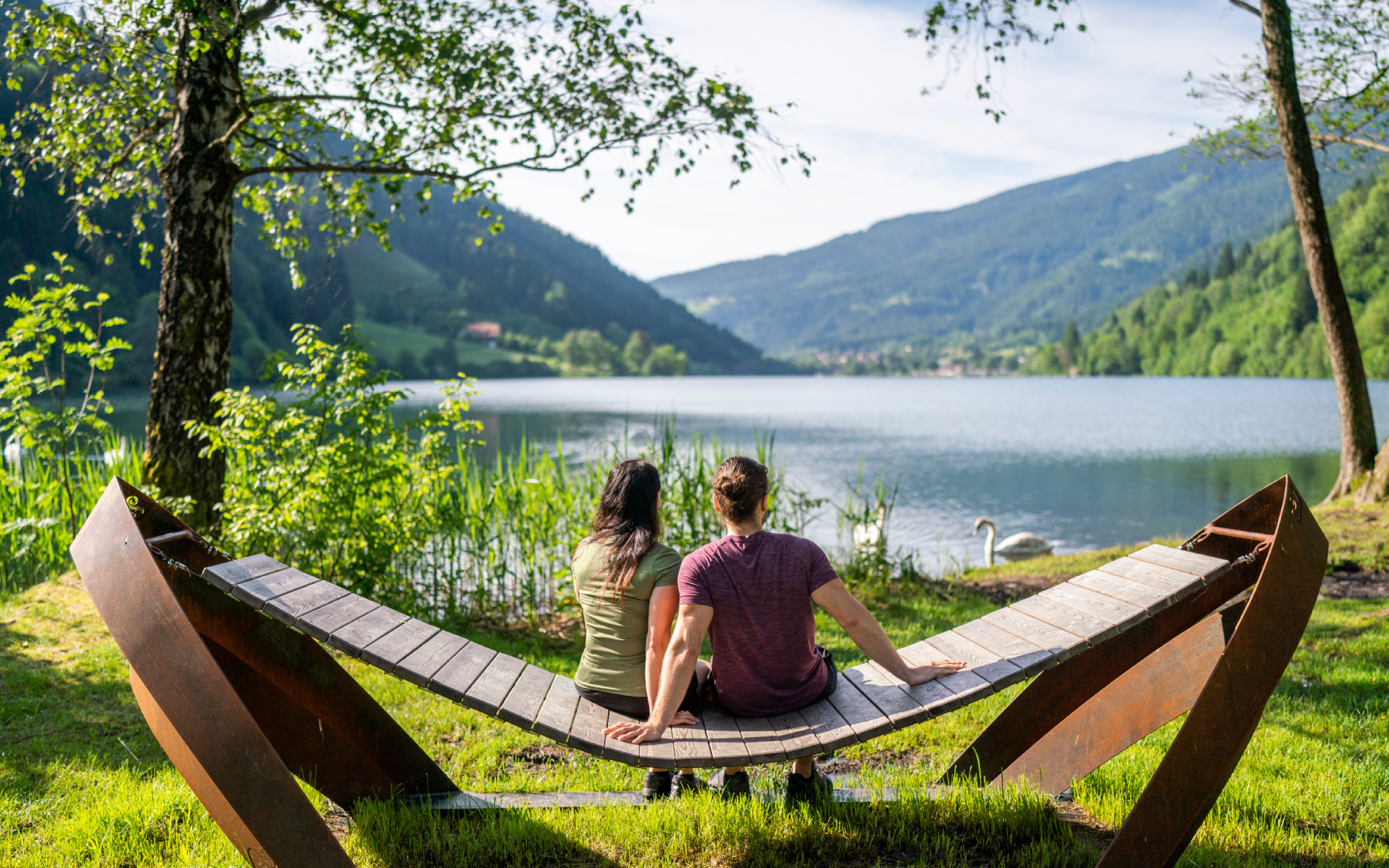 Ein Pärchen sitzt gemütlich auf einer Hängesessel und genießt den schönen Ausblick auf den See und der umliegenden Berglandschaft